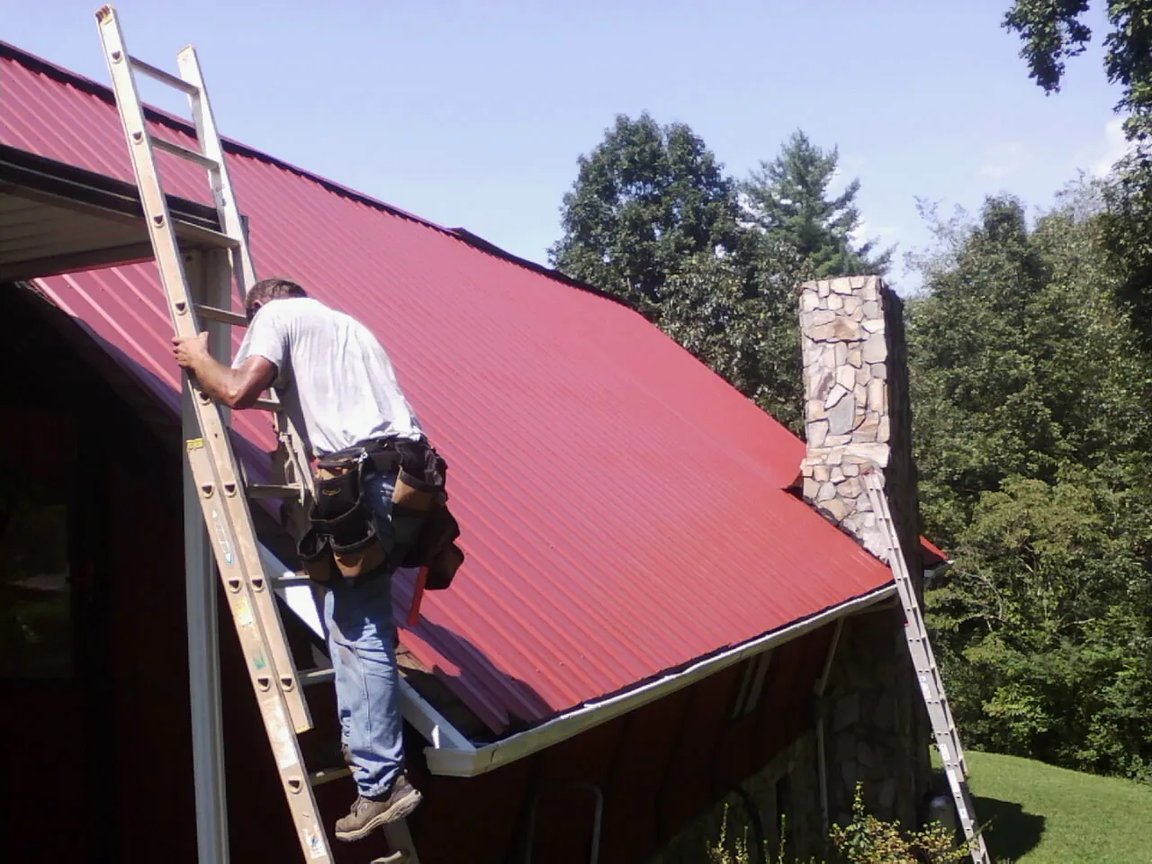 Roofer on a ladder beside a red metal roof and stone chimney in Downers Grove
